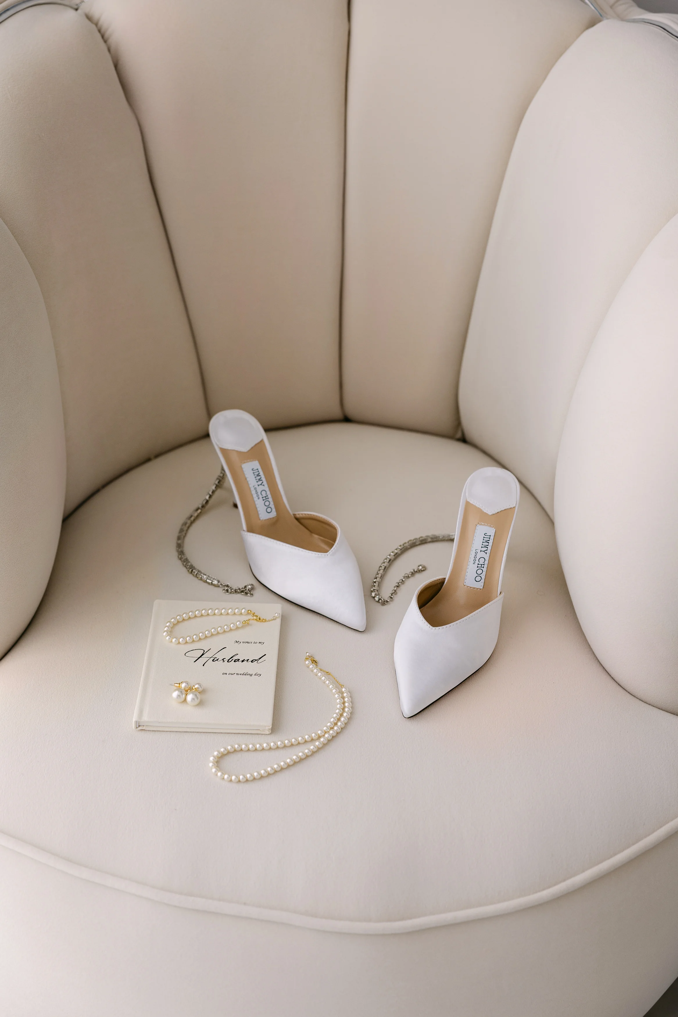 Bridal shoes, pearls, and earrings styled on a cream lounge chair before the wedding.