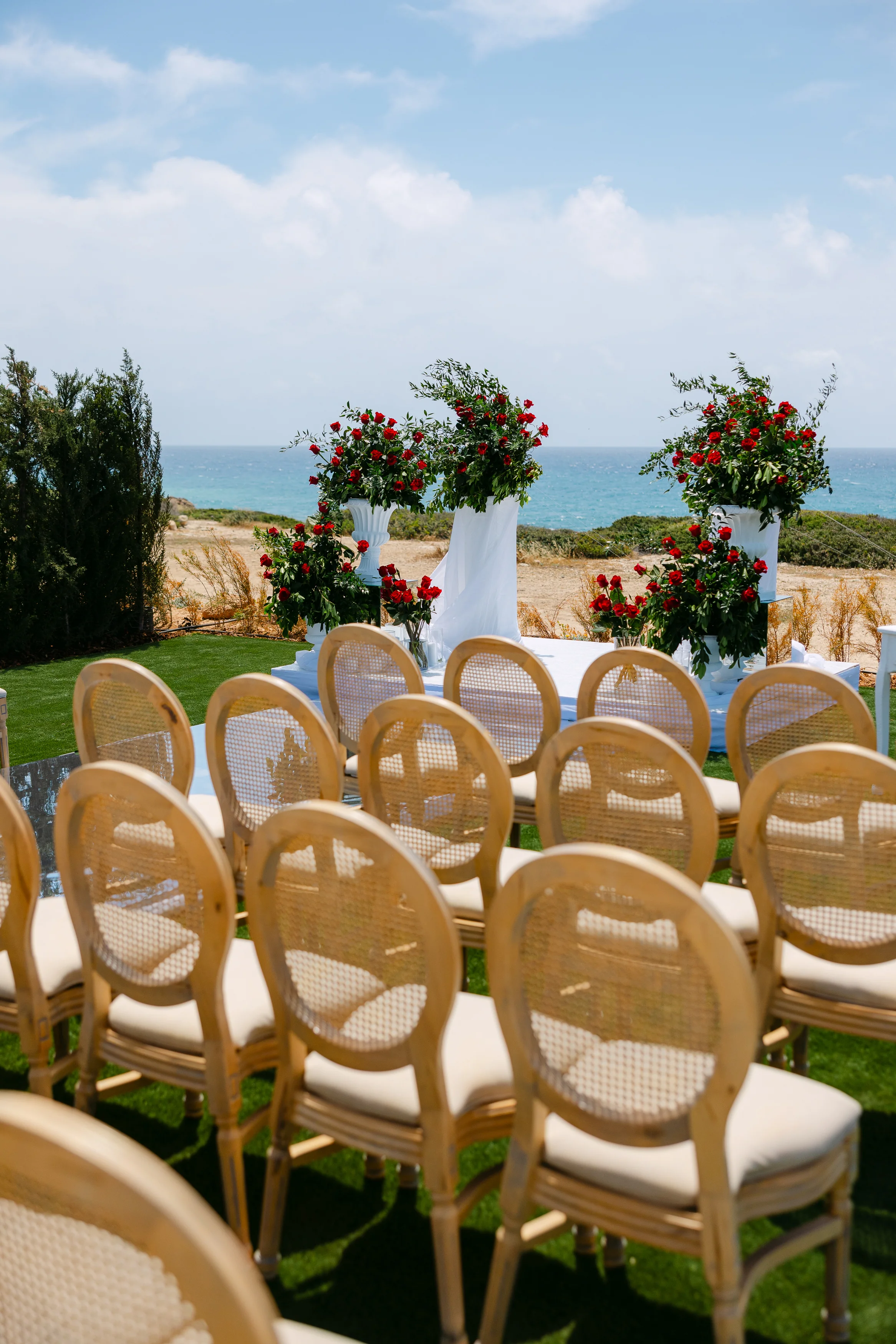 Cane-back ceremony chairs lined up at OZEA with red and white floral arrangements and the sea beyond.