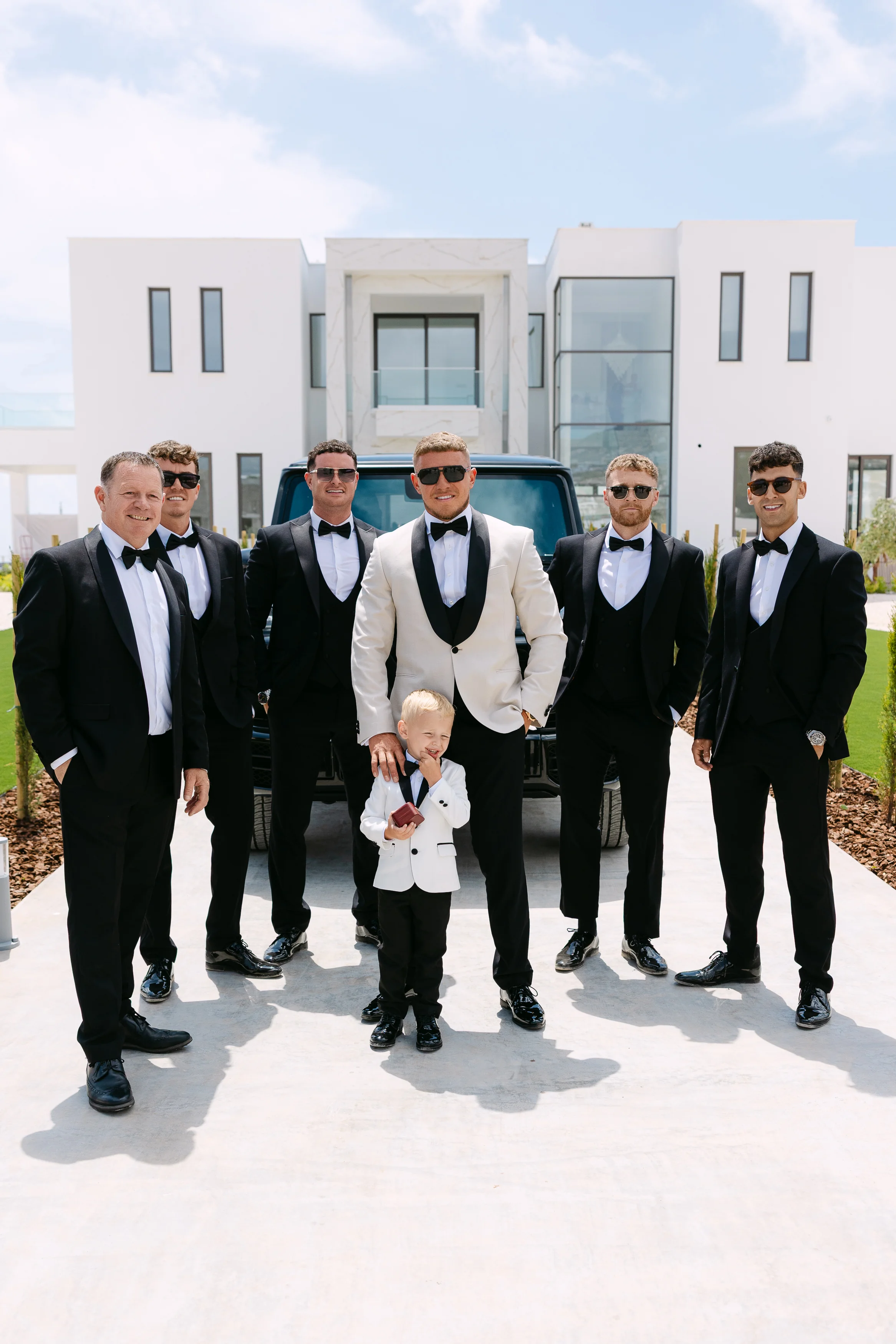 Groom, groomsmen, and young ring bearer photographed in front of the venue with a black Mercedes G-Wagon.