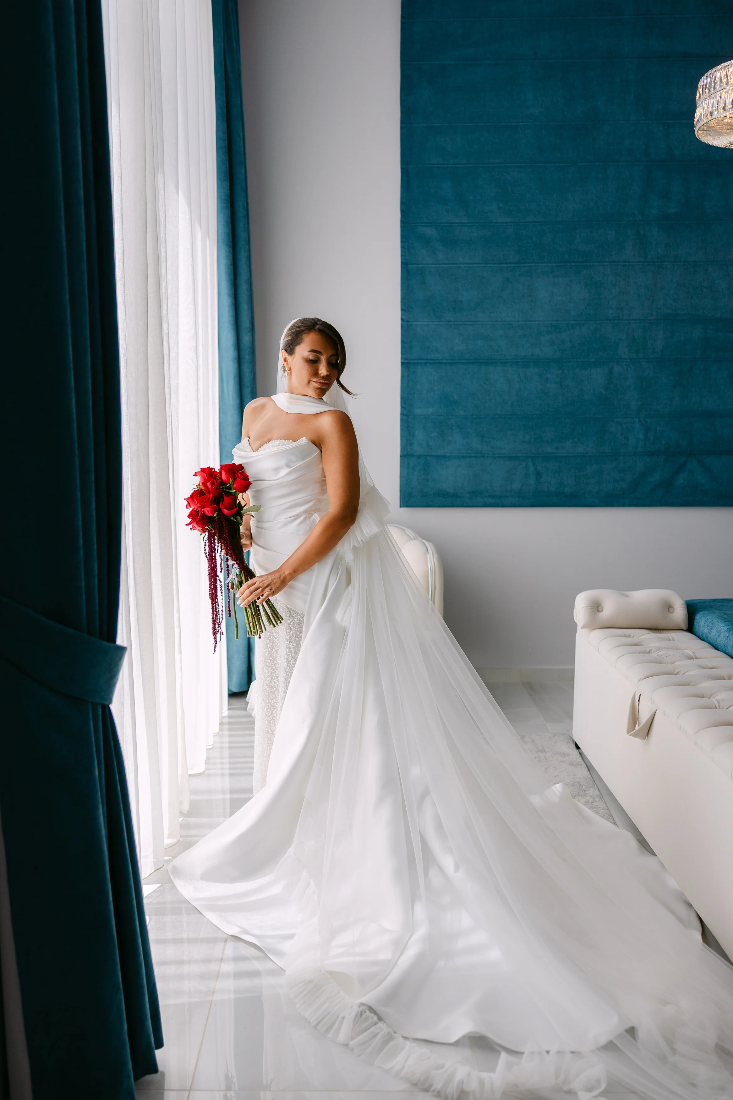 Bride in her wedding gown holding a red bouquet beside the window in the bridal suite at OZEA.