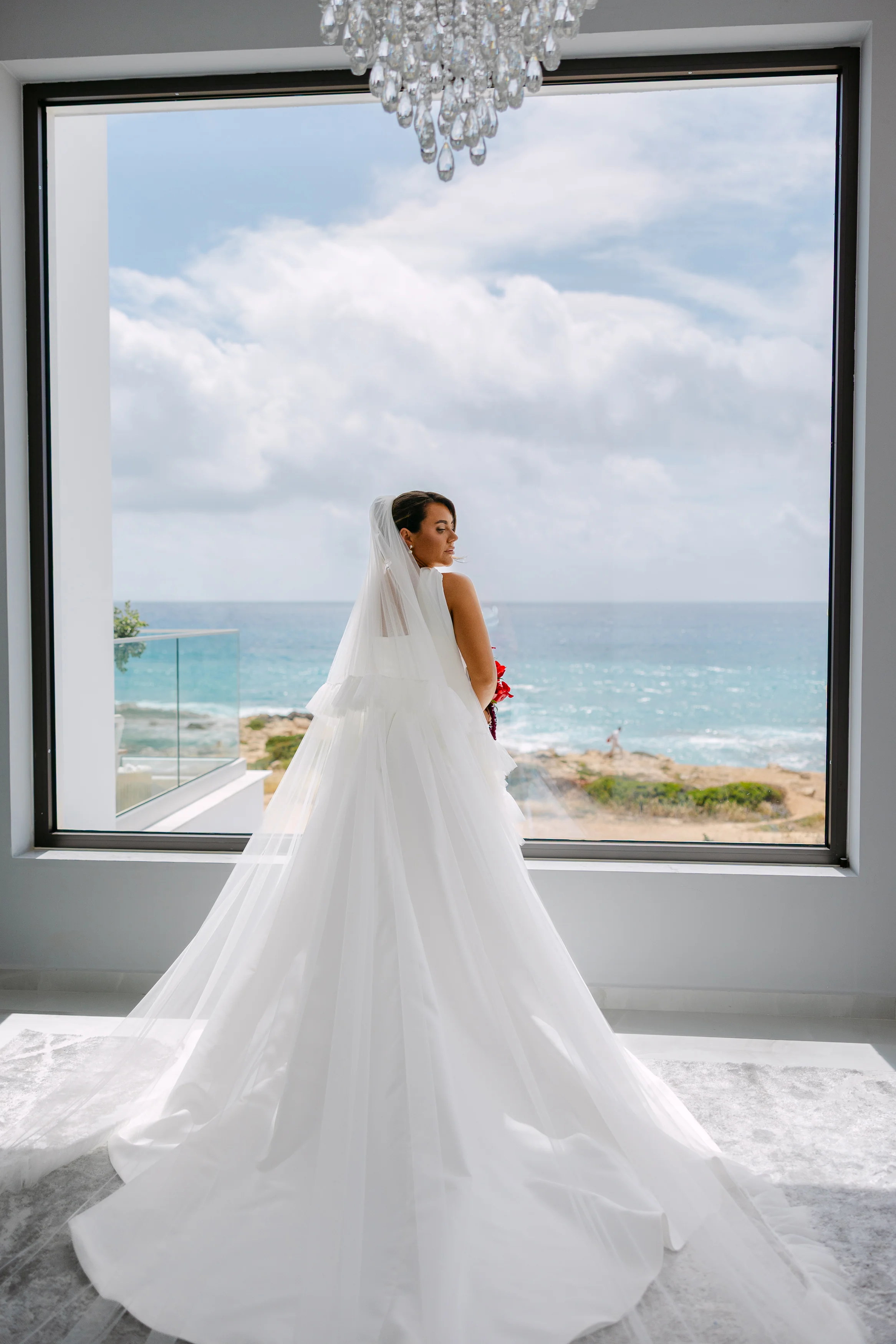 Bride beneath a crystal chandelier facing a panoramic window with the Paphos coastline beyond.