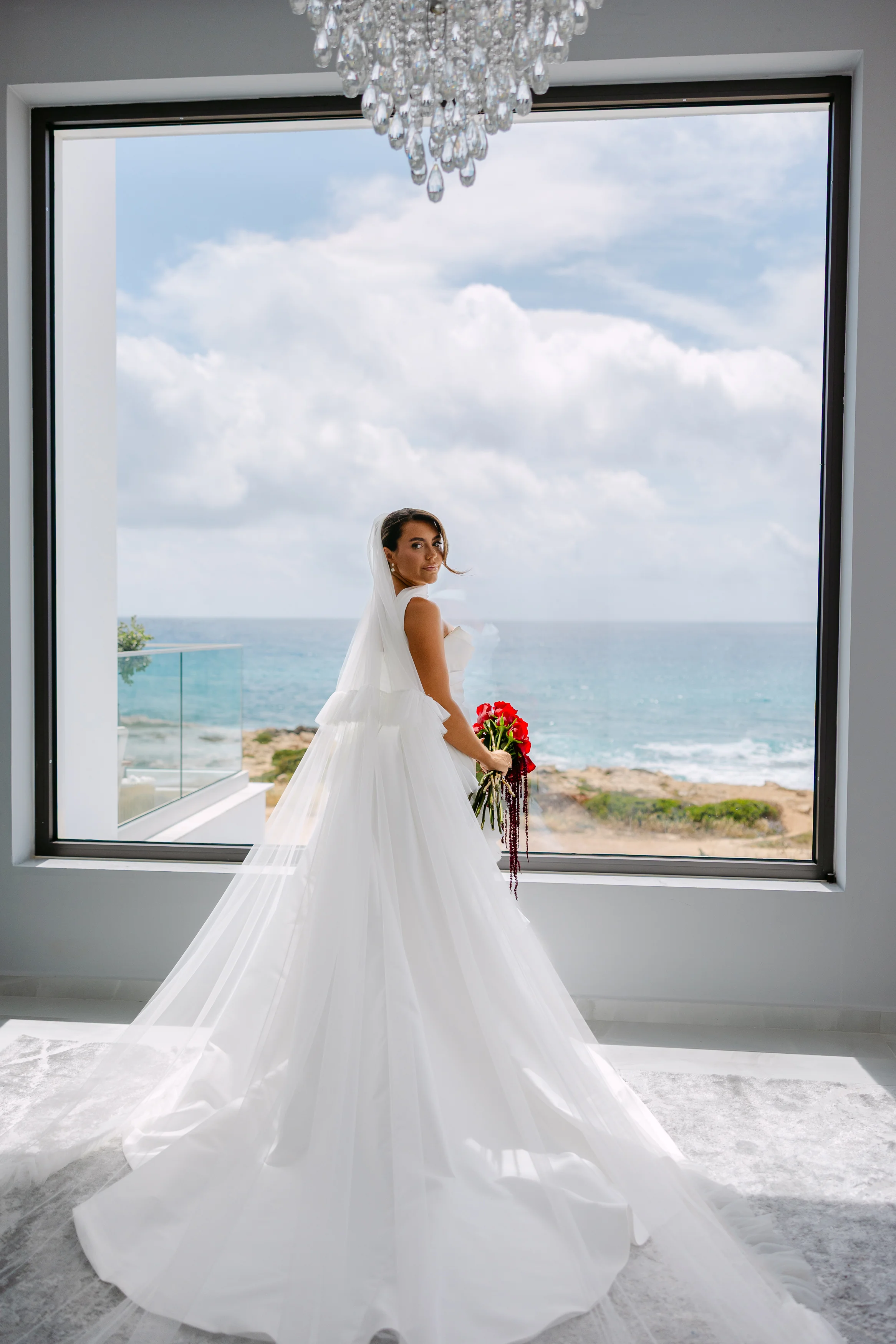 Back view of the bride at the bridal suite’s sea-view window, full train and red bouquet.