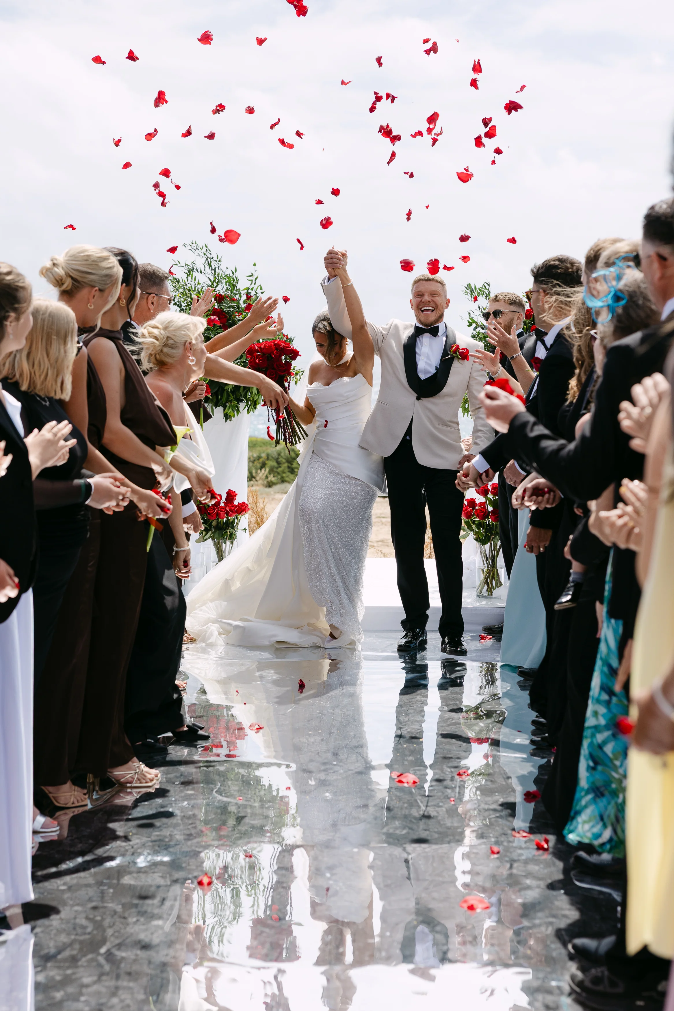 Newlywed couple raising joined hands as red petal confetti falls during their ceremony exit at OZEA.