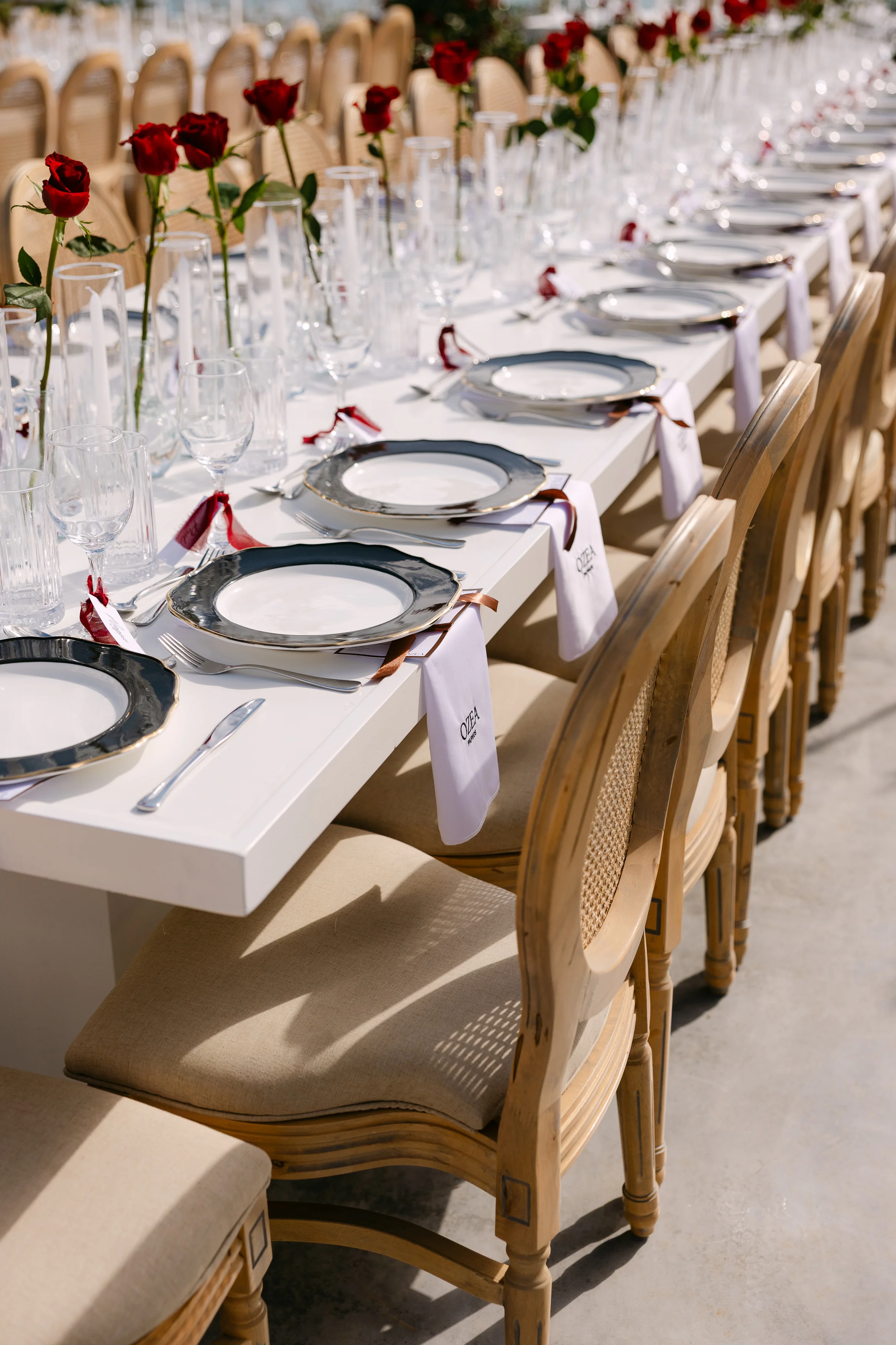 Close view of a long banquet table at OZEA with grey-rimmed plates, red rose buds in glass vessels, and personalised place cards.