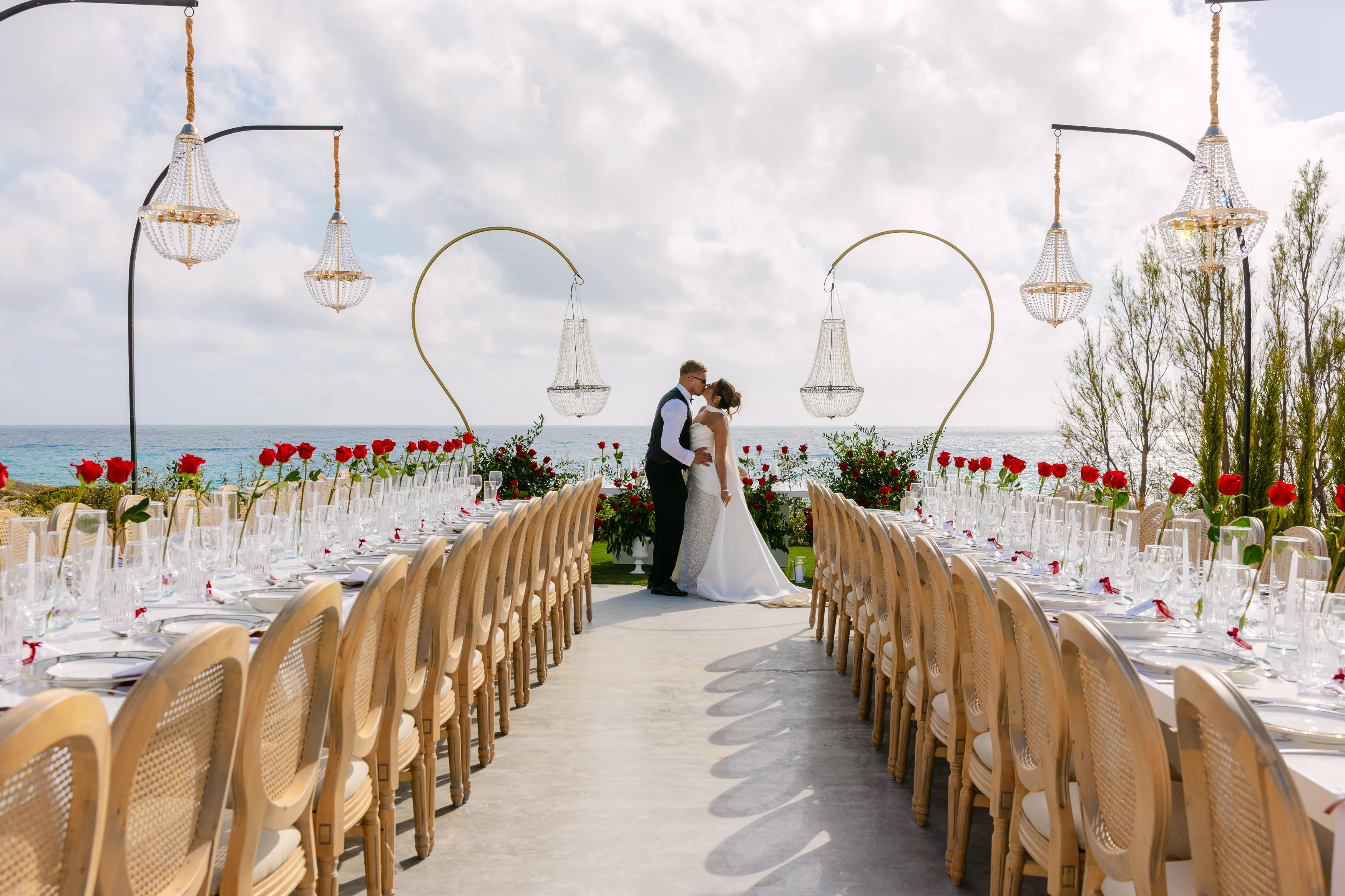 Bride and groom kiss between the long banquet tables at OZEA with chandeliers above and the sea behind.