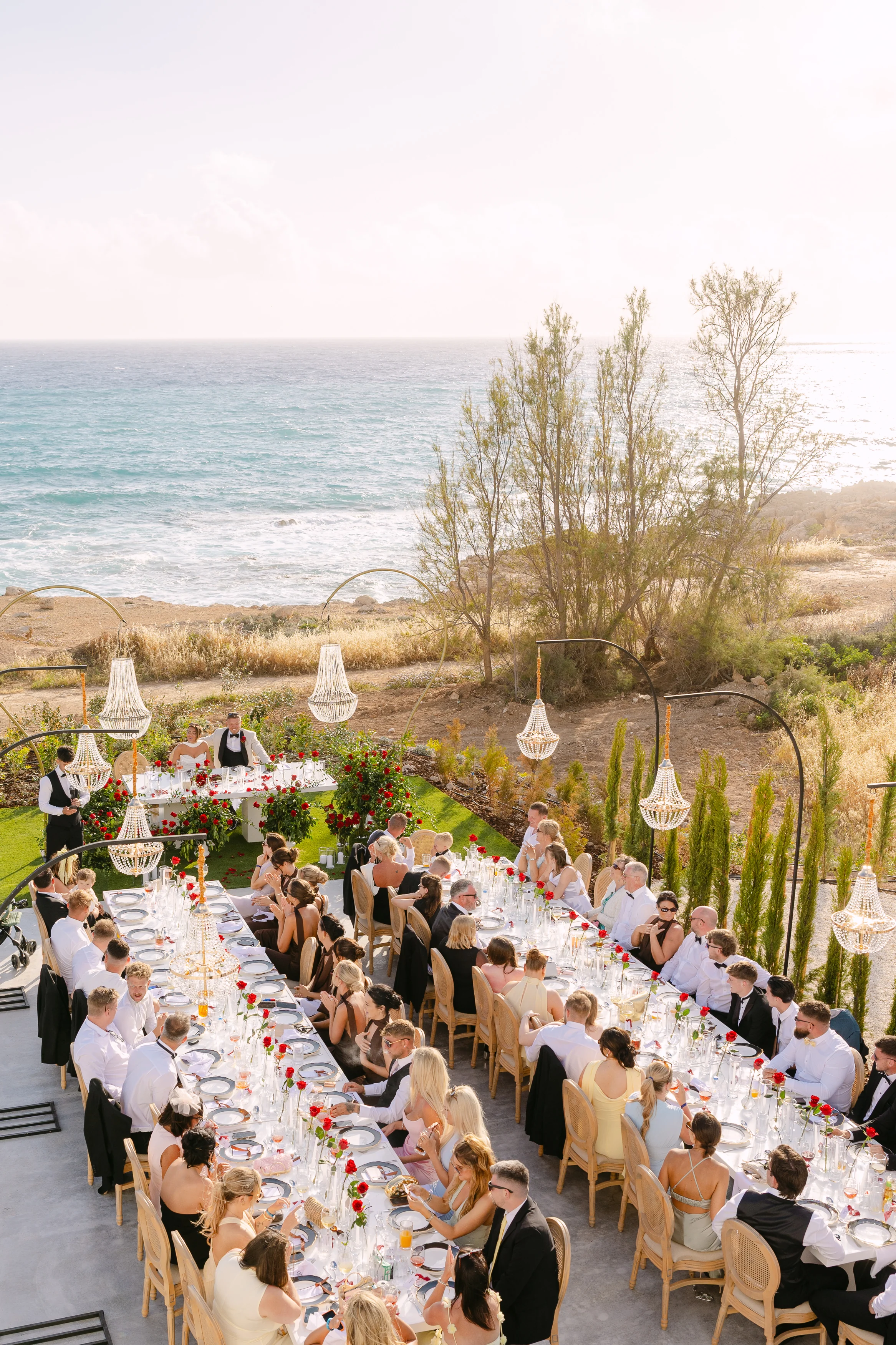 Elevated golden-hour view of a wedding banquet at OZEA with guests seated, chandeliers lit, and the Mediterranean glowing behind.