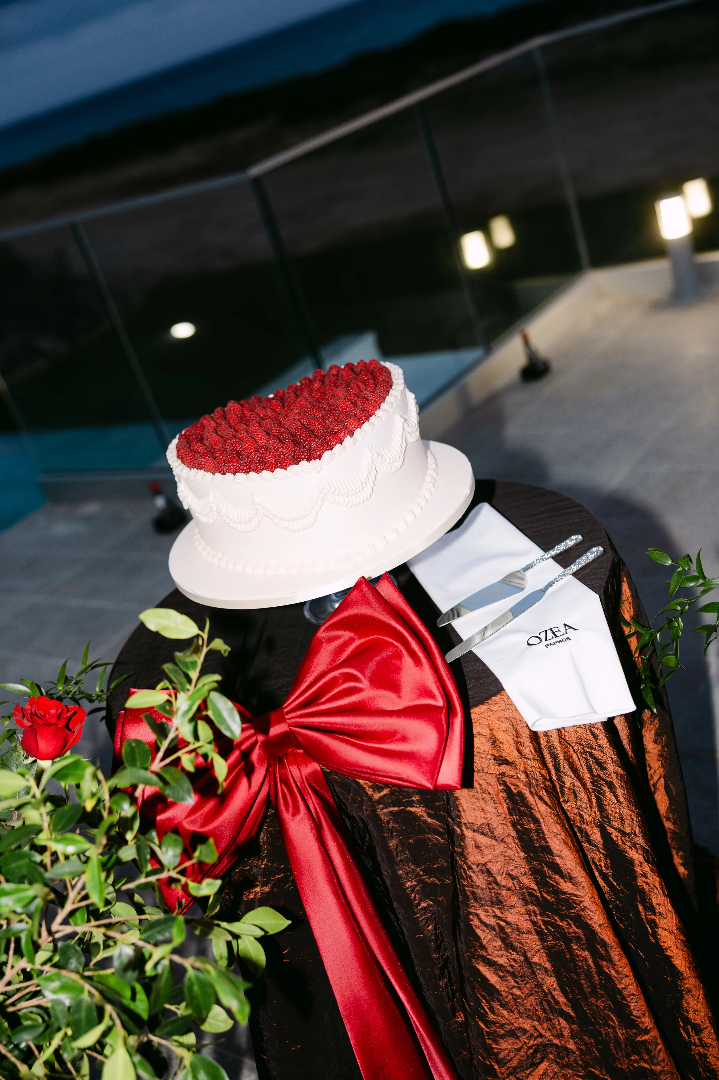 Wedding cake topped with red roses on a black silk-draped table with a wide red bow at the front.