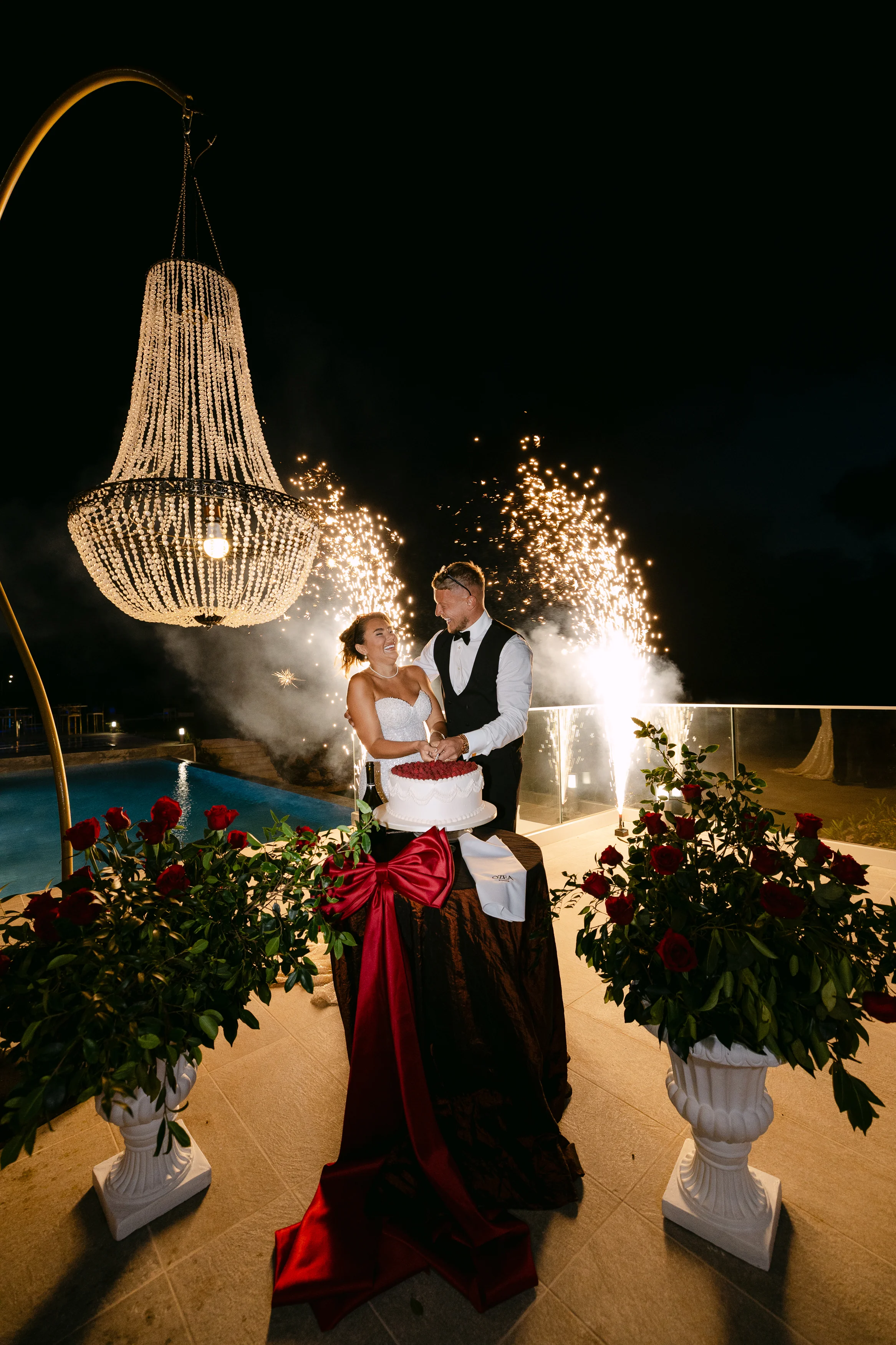 Bride and groom cutting the cake beneath a chandelier at Ozea with sparkler fountains and red roses framing.