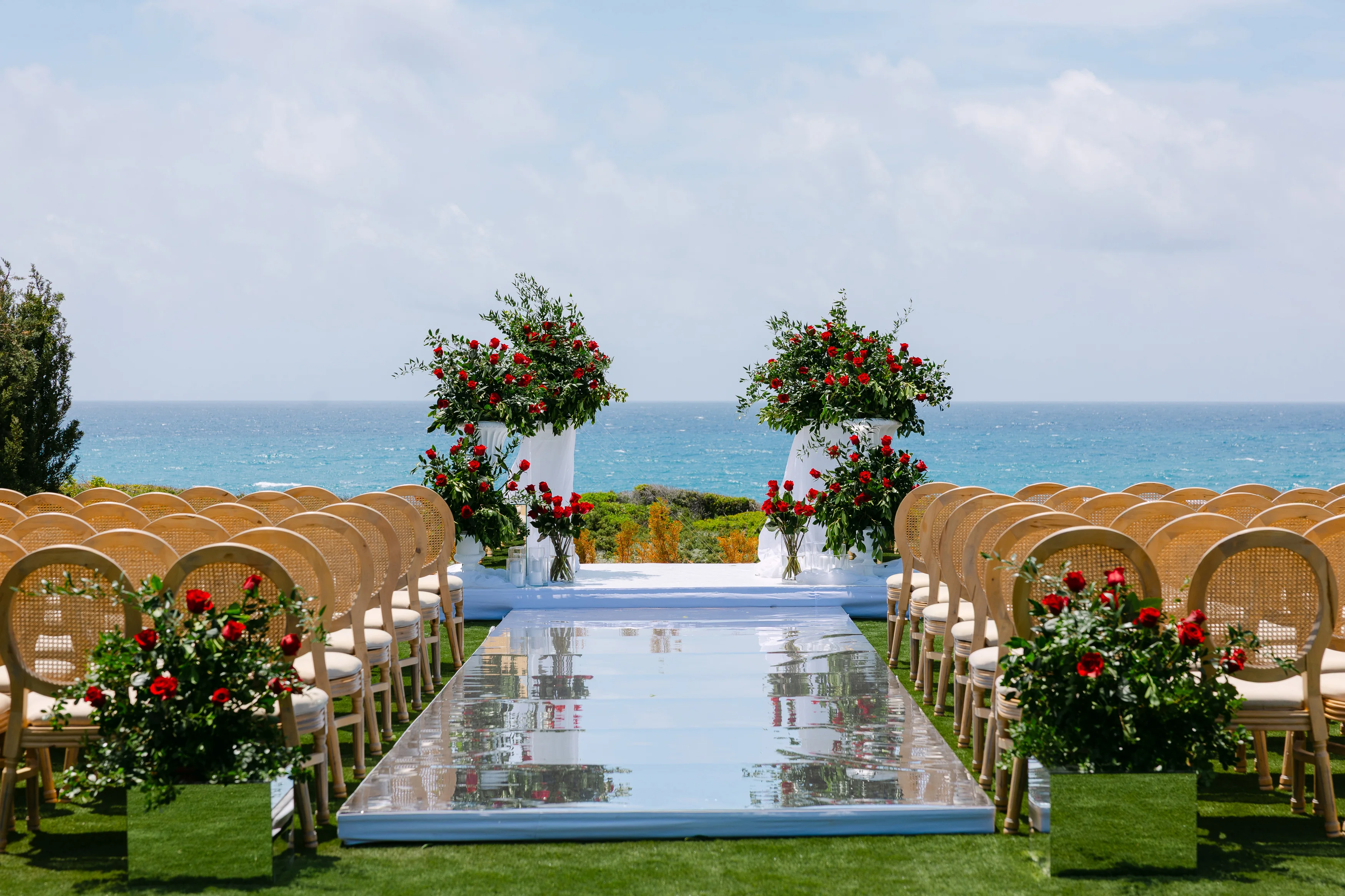 Ceremony seating at Ozea with cane-back chairs and red rose pedestals facing the sea.
