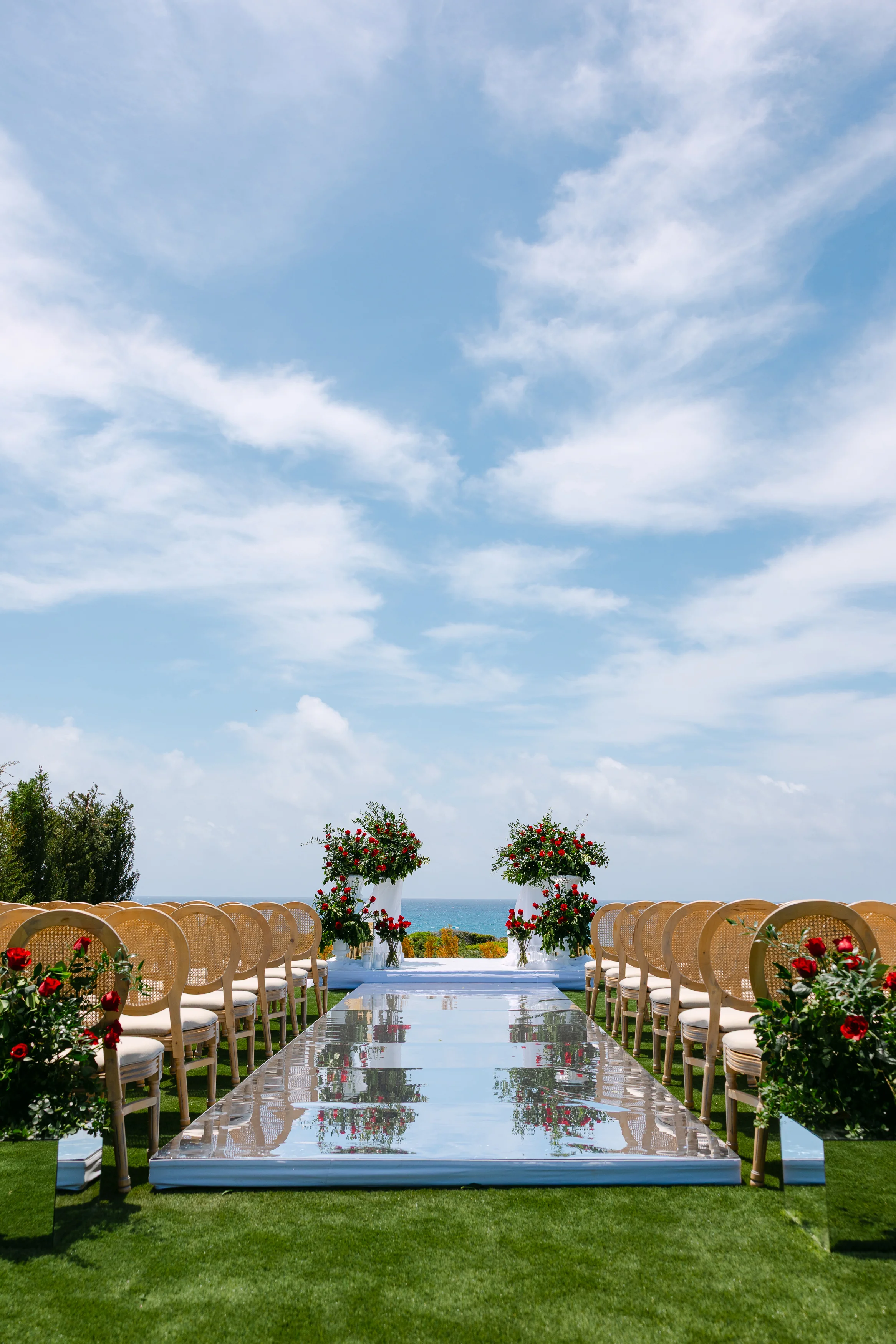 Vertical view down the ceremony aisle at OZEA with floral arrangements, mirrored runway, and the sea ahead.